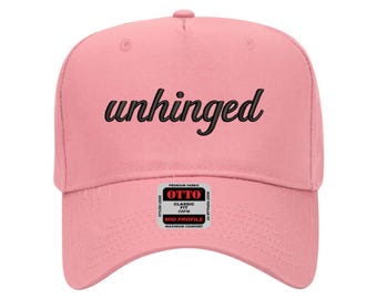 May include: A light pink baseball cap with the word "unhinged" in black script across the front. The cap has a curved brim and a small tag on the side. The cap is made of a soft fabric and has a classic fit.