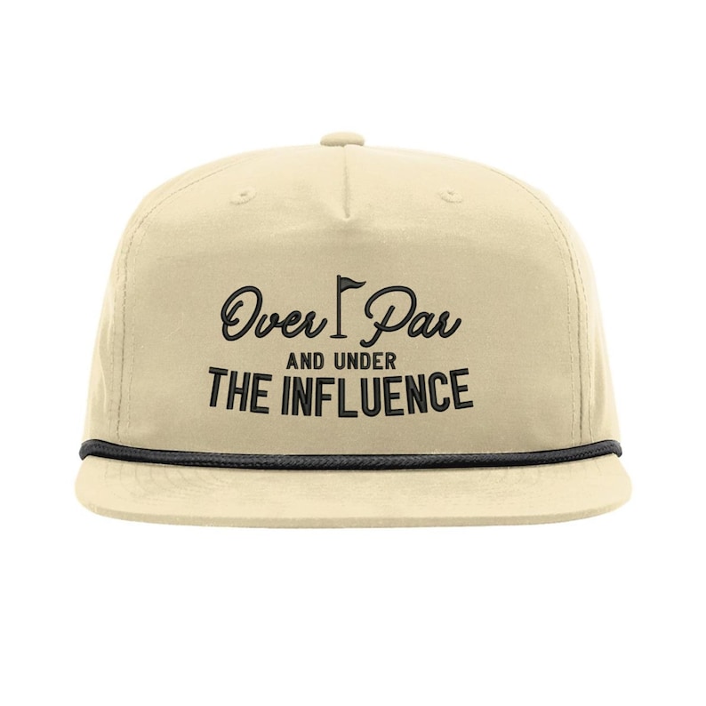 Op de afbeelding: Beige pet met platte klep en een zwart koorddetail langs de rand. De pet heeft de tekst "Over Par" boven "And Under The Influence" in zwarte letters, met een golfvlag.