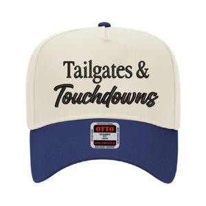 May include: A baseball cap with a cream-colored crown and a navy blue brim. The words "Tailgates & Touchdowns" are embroidered in black script across the front. The cap has a structured fit and a small label.
