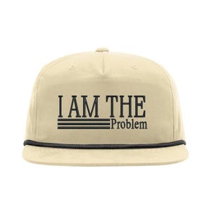 以下が含まれることがあります： 黒色のロープトリムと「I AM THE Problem」という黒色の刺繍文字が施されたベージュのベースボールキャップです。