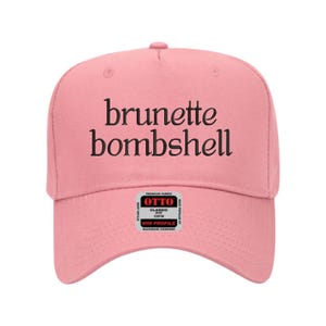 May include: A pink baseball cap with the words "brunette bombshell" in black script across the front. The cap has a curved brim and a small tag on the side that reads "OTTO" and "MID PROFILE".