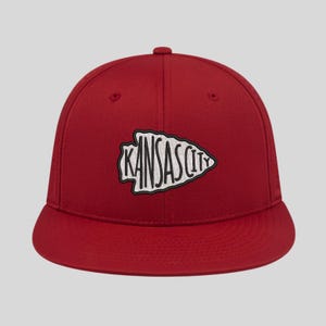 Puede incluir: Gorra de béisbol roja con un diseño de punta de flecha blanca con el texto "KANSAS CITY" en negro.