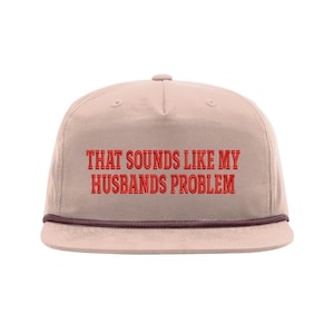 May include: A light tan, flat-brimmed cap with the embroidered text "THAT SOUNDS LIKE MY HUSBANDS PROBLEM" in red. The cap has a brown rope detail along the brim and a button on top.