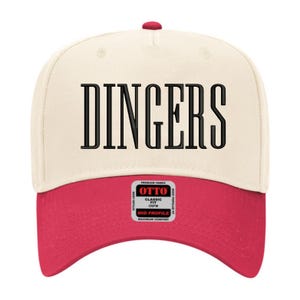 May include: A baseball cap with a cream-colored crown and a red brim. The word "DINGERS" is printed in large, black, block letters across the front. The cap has a curved brim and a red button on top.