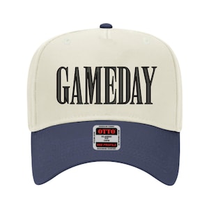 May include: A baseball cap with a cream-colored crown and a navy blue brim. The word "GAMEDAY" is embroidered in large, black letters across the front. The cap has a curved bill and a small navy blue button on top.