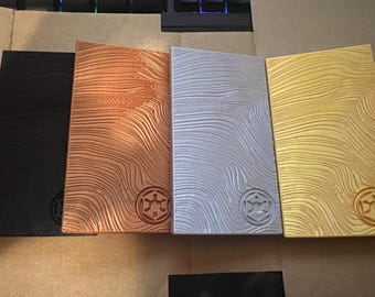 May include: Four rectangular metal panels in black, copper, silver, and gold. Each panel has a textured, wood-grain-like pattern and a circular emblem in the lower right corner. The panels are arranged side by side.