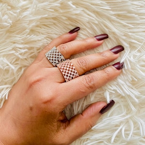 May include: Two wide band rings, one silver-toned and one rose gold-toned, each encrusted with small, sparkling stones. The rings are displayed on a fluffy, white surface, creating a contrast with the jewelry's shine.