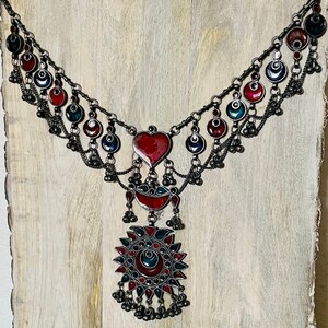 May include: A silver necklace with red and blue enamel accents. The necklace features a heart-shaped pendant and a large circular pendant with a sunburst design.