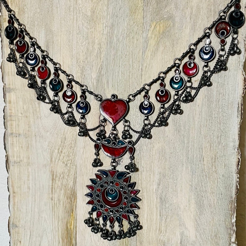 May include: A silver necklace with red and blue enamel accents. The necklace features a heart-shaped pendant and a large circular pendant with a sunburst design.