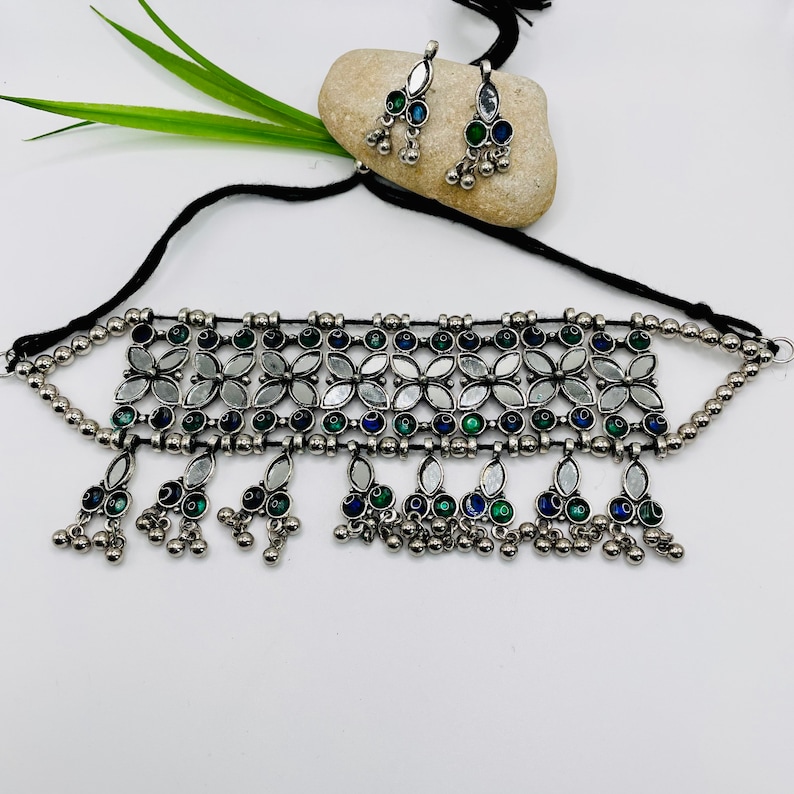 May include: A silver-toned necklace and matching earrings set. The necklace features a floral design with mirrored accents and blue and green details. The earrings and necklace have dangling elements with small silver beads. The set is displayed on a white background.