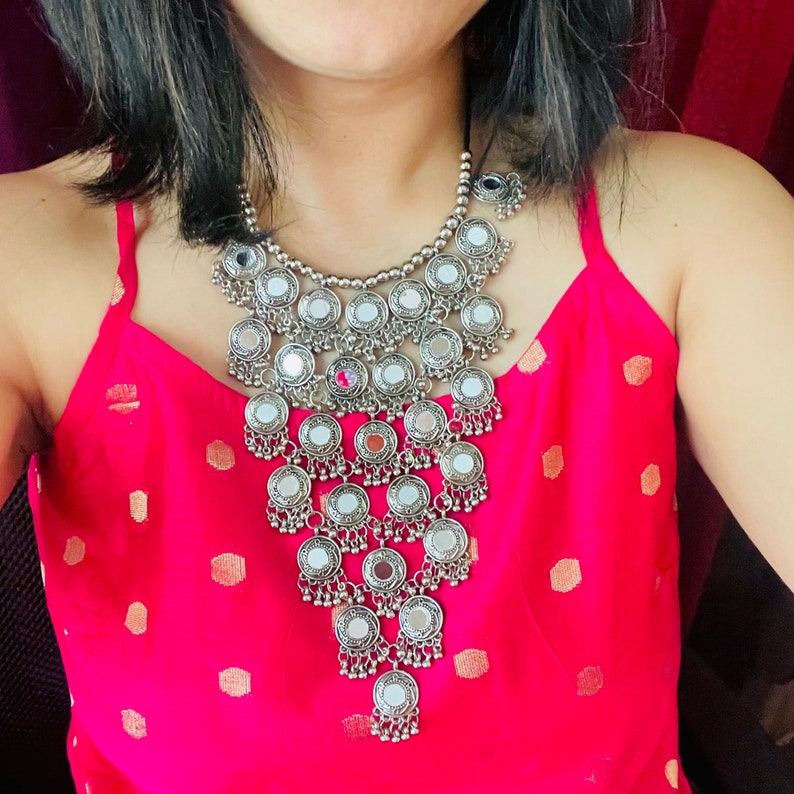 May include: A silver-toned statement necklace with multiple circular mirrored pendants. The necklace is layered and hangs over a bright pink top. The design includes small dangling charms at the bottom of the pendants. The background is a solid color.