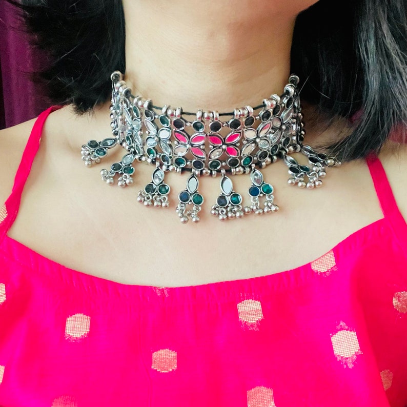 May include: Silver-toned choker necklace with a floral design featuring pink, green, and clear accents. The necklace has dangling pendants with small silver beads. The model is wearing a bright pink top.