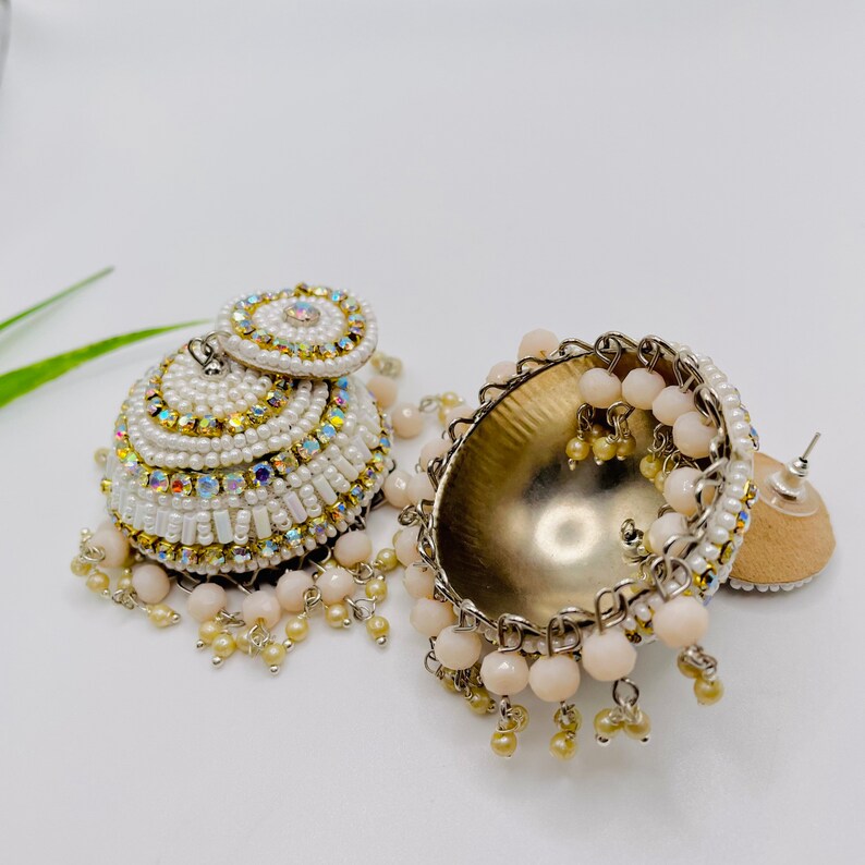 K&ouml;nnte beinhalten: Ein Paar wei&szlig;e und goldene Perlenohrringe mit einem baumelnden Design. Die Ohrringe haben einen runden, perlenbesetzten oberen Teil mit einer silbernen R&uuml;ckseite und einem langen, perlenbesetzten Fransen.