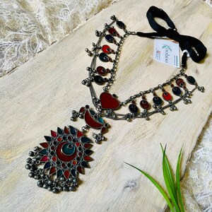 May include: A silver necklace with a large, colorful pendant featuring a sunburst design. The pendant is made of glass and metal and has red, blue, and green accents. The necklace is also adorned with smaller, circular pendants with red, blue, and green accents. The necklace is tied with a black ribbon.