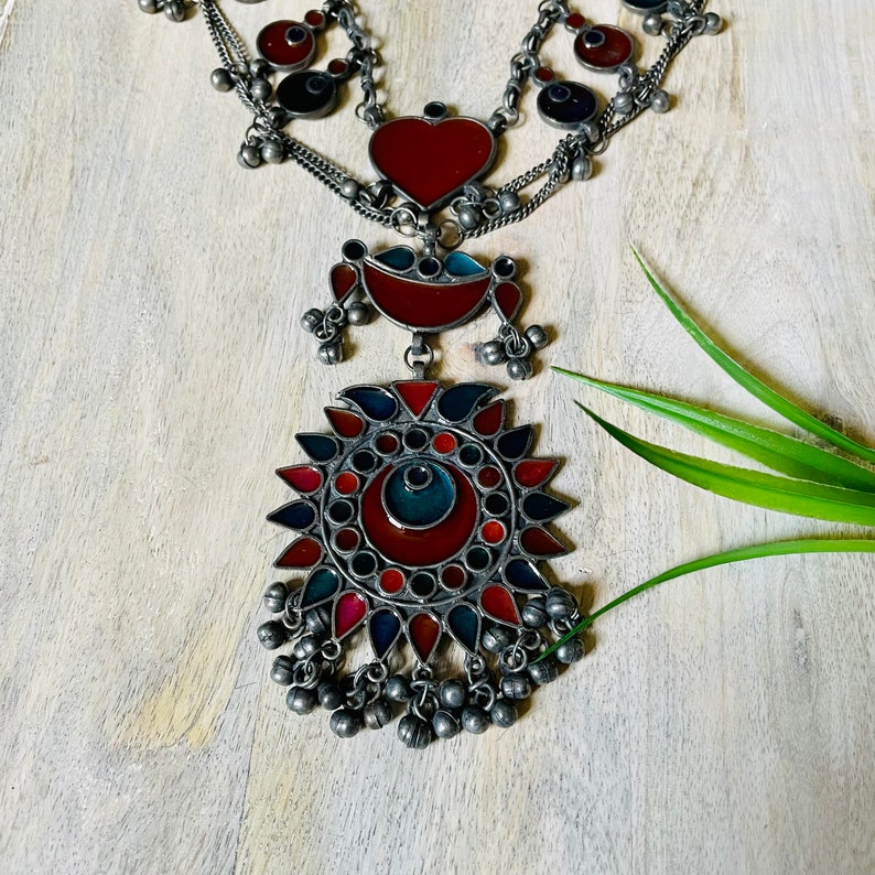 May include: A silver necklace with multiple chains and a large pendant featuring a red and blue enamel design. The pendant is surrounded by smaller, round enamel pieces in red, blue, and green. The necklace is adorned with small silver bells.
