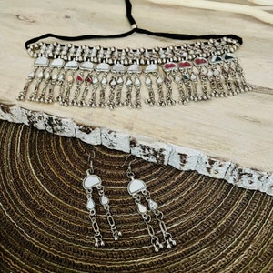May include: A silver-toned choker necklace and matching earrings set. The necklace has a black cord and a row of small, round beads. The earrings and necklace feature a series of dangling, teardrop-shaped charms. The jewelry is displayed on a textured, wooden surface.