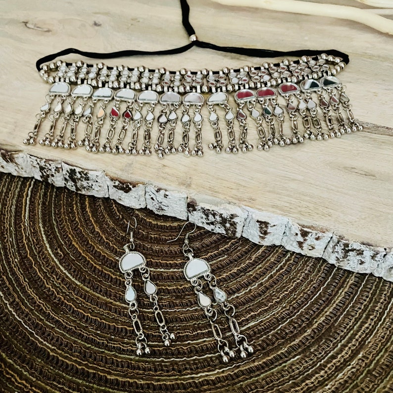 May include: A silver-toned choker necklace and matching earrings set. The necklace has a black cord and a row of small, round beads. The earrings and necklace feature a series of dangling, teardrop-shaped charms. The jewelry is displayed on a textured, wooden surface.