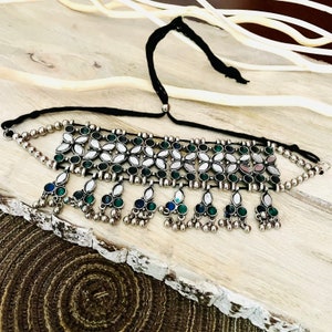 May include: Silver-toned choker necklace with a black cord. The necklace features a floral design with green and white accents, and dangling teardrop-shaped charms with green and blue stones. The necklace is displayed on a wooden surface.