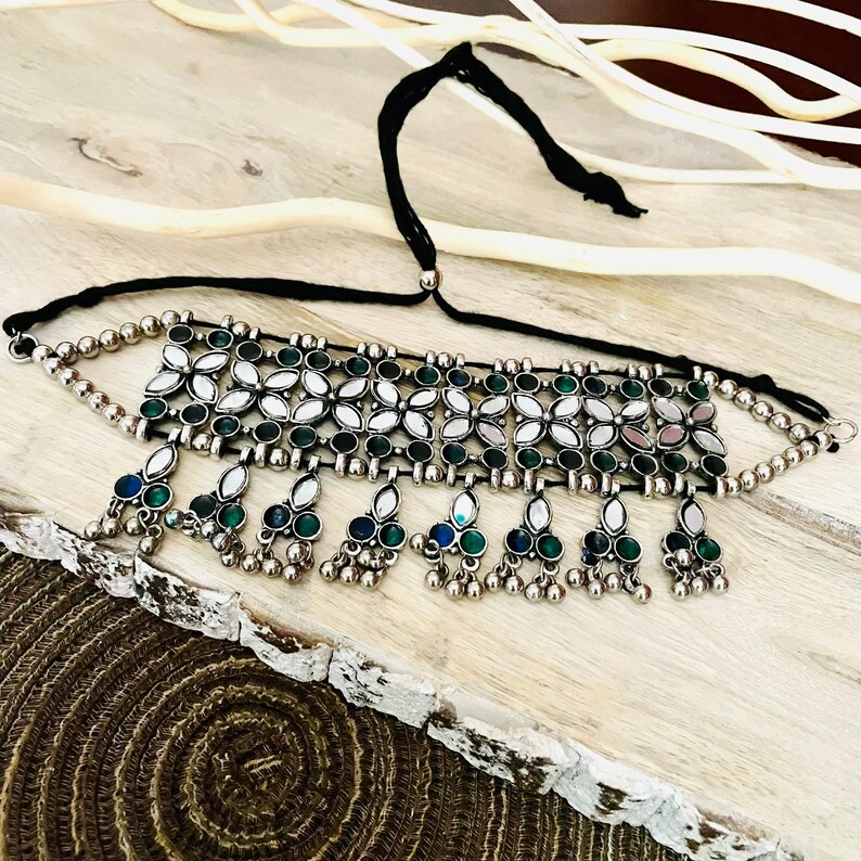 May include: Silver-toned choker necklace with a black cord. The necklace features a floral design with green and white accents, and dangling teardrop-shaped charms with green and blue stones. The necklace is displayed on a wooden surface.