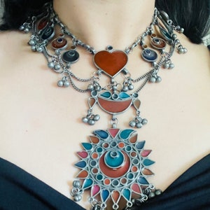 May include: A silver necklace with multiple layers of chains and colorful glass pendants. The pendants are in the shape of a heart, a crescent moon, and a sun with a circular design. The necklace has a bohemian style.