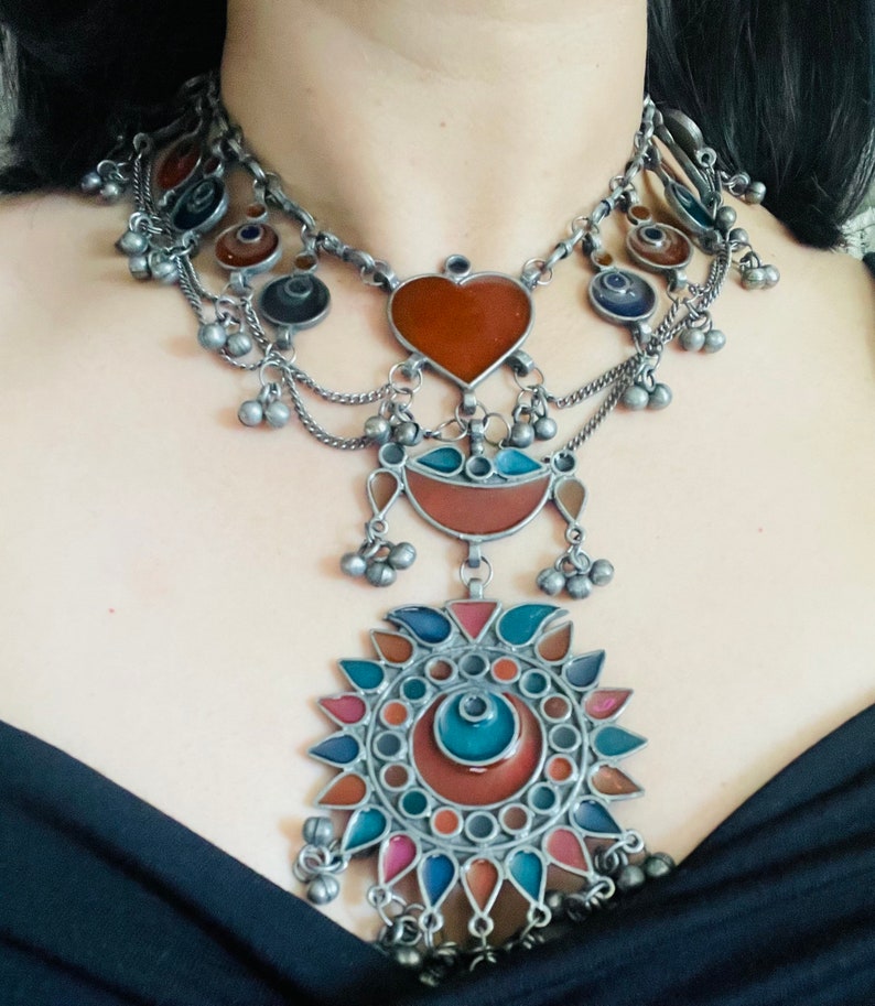 May include: A silver necklace with multiple layers of chains and colorful glass pendants. The pendants are in the shape of a heart, a crescent moon, and a sun with a circular design. The necklace has a bohemian style.