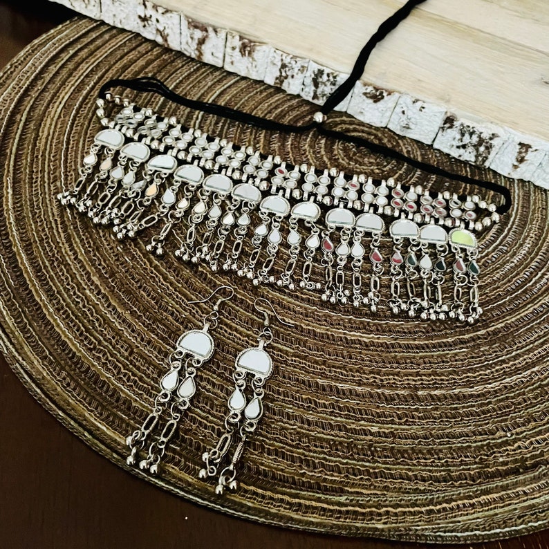 May include: A silver-toned necklace and matching earrings set. The necklace features a choker style with a black cord and dangling mirrored accents. The earrings have a similar design, with mirrored elements and dangling chains. The jewelry is displayed on a brown woven mat.