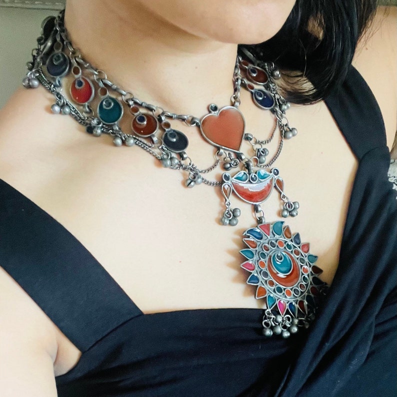 May include: A silver necklace with colorful enamel accents. The necklace features a heart-shaped pendant, a crescent moon pendant, and a large circular pendant with a sunburst design.