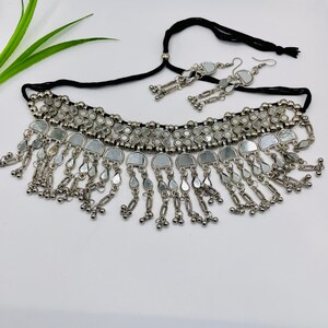 May include: A silver-toned necklace and matching earrings set. The necklace features a black cord and is adorned with small, round mirrors and dangling teardrop-shaped accents. The earrings mirror the necklace design, creating a cohesive jewelry set.