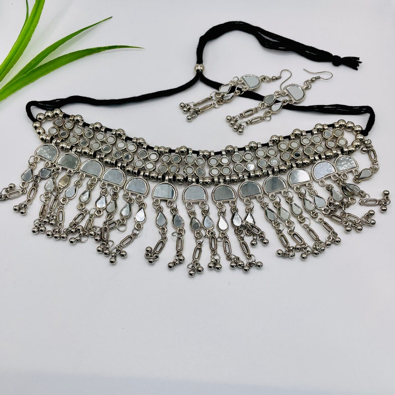 May include: A silver-toned necklace and matching earrings set. The necklace features a black cord and is adorned with small, round mirrors and dangling teardrop-shaped accents. The earrings mirror the necklace design, creating a cohesive jewelry set.