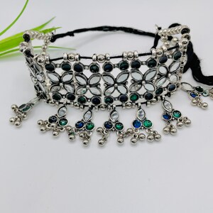 May include: A silver-toned choker necklace with a floral design. The necklace features clear, black, blue, and green gemstones. Dangling accents with small silver beads and gemstones hang from the bottom of the choker. The necklace is on a black cord.