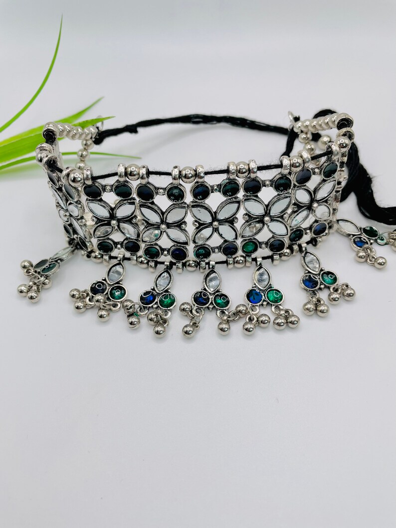 May include: A silver-toned choker necklace with a floral design. The necklace features clear, black, blue, and green gemstones. Dangling accents with small silver beads and gemstones hang from the bottom of the choker. The necklace is on a black cord.