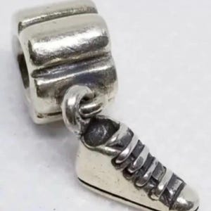 Puede incluir: Un charm de plata con una cuenta rectangular y un pequeño colgante de zapatilla deportiva. La zapatilla tiene cordones detallados y una suela texturizada. El charm está diseñado para ser añadido a una pulsera o collar.