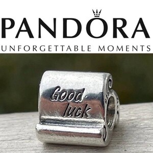 以下が含まれることがあります： 「Good luck」の文字が刻まれたシルバーのパンドラチャーム。チャームは長方形で、縁が丸められており、ブレスレットに通すための穴があります。背景にはパンドラのロゴと「Unforgettable Moments」の文字があります。