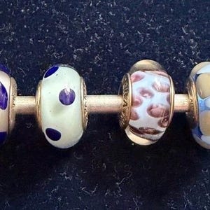 以下が含まれることがあります： 銀色の棒に4つの装飾ビーズ。ビーズは、青い葉の模様、白と青のドット、茶色と白の斑点、青と金のアクセントの幾何学模様など、さまざまなデザインが特徴です。ビーズはおそらくジュエリー作りのためです。