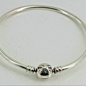 Puede incluir: Una pulsera de plata con un cierre decorativo. La pulsera tiene un diseño simple y circular con un acabado liso y pulido. El cierre presenta un dije redondo y detallado con un diseño intrincado.