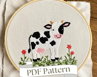 Könnte beinhalten: Gesticktes Kuhdesign in einem Holzreifen. Die Kuh ist weiß mit schwarzen Flecken, rosa Euter und Maul sowie kleinen Hörnern. Rote Blumen und grünes Gras sind unter der Kuh gestickt. Der Text "PDF Pattern" steht auf einem Banner.