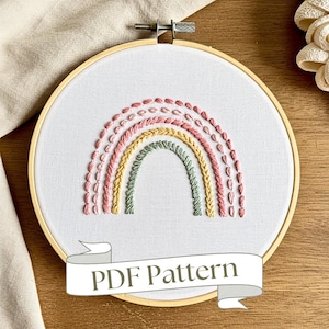 May include: A PDF pattern for a colorful rainbow embroidery hoop art project. The rainbow is made up of pink, yellow, and green thread. The hoop is wooden and has a white fabric background.