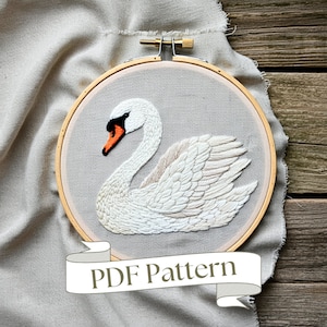 May include: Embroidered swan design in a wooden hoop. The swan features white and beige thread with an orange beak and black accents. The background is a light gray fabric. The text "PDF Pattern" is on a white banner at the bottom.