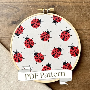 May include: A round embroidery hoop featuring a cream-colored fabric with a repeating pattern of red and black ladybugs. The ladybugs have black heads and spots. The text "PDF Pattern" is on a white banner at the bottom.