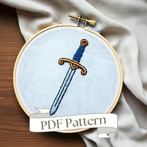 May include: Embroidered sword design on a white fabric stretched in a wooden embroidery hoop. The sword is blue and gold with a gold handle. The hoop is on a wooden surface. The text "PDF Pattern" is on a white ribbon.