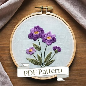 May include: A purple flower embroidery pattern in a wooden embroidery hoop. The pattern features three purple flowers with yellow centres and green leaves. The hoop has a white fabric background and a wooden frame. The text "PDF Pattern" is displayed on a white ribbon.
