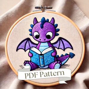 Puede incluir: Un dragón morado con alas azules está sentado y leyendo un libro azul. El dragón tiene ojos grandes y una expresión linda. La imagen está en un aro de bordado de madera. El texto "PDF Pattern" está en una pancarta blanca debajo del aro.