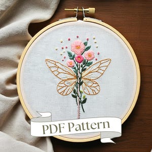 Könnte beinhalten: Stickrahmen mit einem Blumenmuster, das einen Schmetterling mit zarten Flügeln und rosa Blüten zeigt. Der Rahmen ist aus Holz und hat einen weißen Stoffhintergrund. Der Text "PDF Pattern" ist auf einem Band unter dem Rahmen abgebildet.