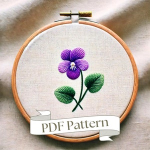 May include: A purple and green embroidered flower design on a white fabric stretched in a wooden embroidery hoop. The hoop has a wooden screw on the top. The text "PDF Pattern" is on a white ribbon banner below the hoop.