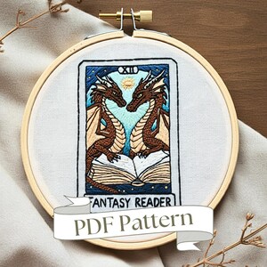 Op de afbeelding: Borduurkunst in een hoepel met een tarotkaart met twee draken die een boek lezen. De kaart heeft de titel "Fantasy Reader" en heeft een blauwe en witte achtergrond met sterren. De hoepel is gemaakt van hout en heeft een natuurlijke afwerking.