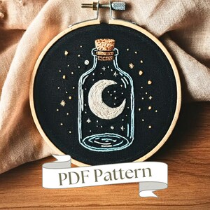 Puede incluir: Un aro de bordado negro con una luna creciente blanca dentro de una botella de vidrio llena de estrellas blancas sobre un fondo negro. La botella tiene un tapón de corcho. El aro tiene una cinta blanca con el texto "PDF Pattern" en ella.