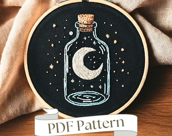 Peut inclure: Un cerceau de broderie noir avec une lune croissante blanche à l'intérieur d'une bouteille en verre remplie d'étoiles blanches sur un fond noir. La bouteille a un bouchon en liège. Le cerceau a un ruban blanc avec le texte "PDF Pattern" dessus.