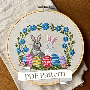 May include: Embroidered hoop art featuring two bunnies surrounded by a wreath of blue flowers and colorful Easter eggs. The hoop has a wooden frame and the text "PDF Pattern" is displayed on a banner below the hoop.