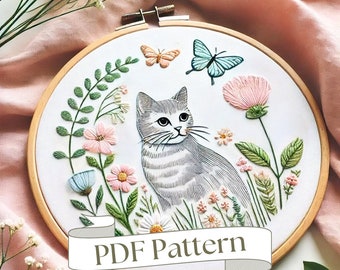 May include: Embroidered hoop art featuring a grey tabby cat sitting in a field of flowers. The cat is surrounded by pink, white and blue flowers, and there are two butterflies flying above. The hoop is made of wood and has a white fabric background. The text "PDF Pattern" is displayed on a banner below the hoop.