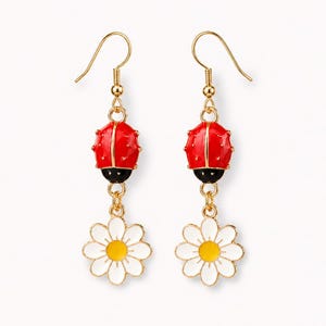 May include: A pair of gold-tone drop earrings. Each earring has a red ladybird charm with black detailing, linked to a white daisy with a yellow centre. The earrings have a hook fastening.